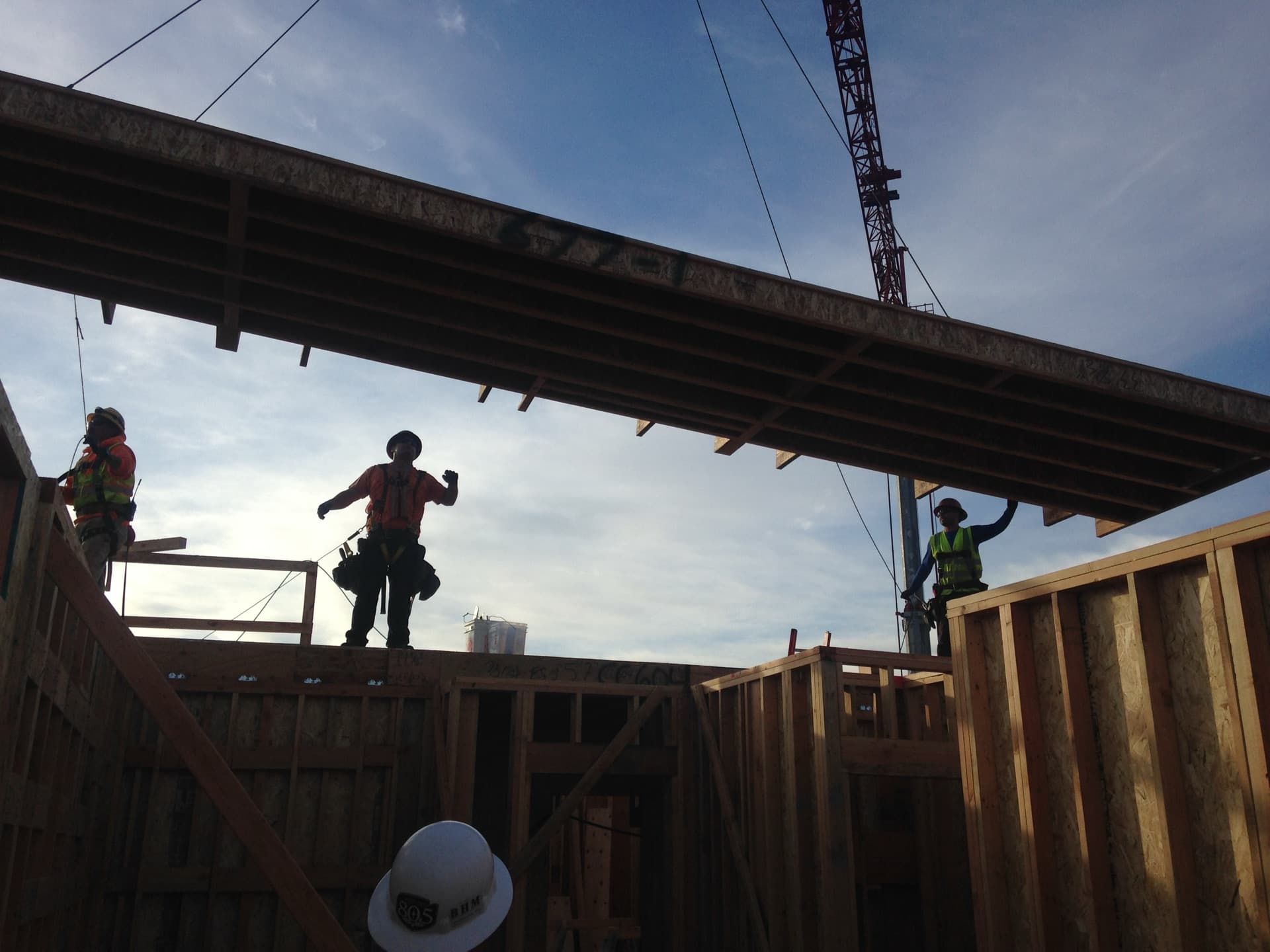 Construction workers silhouetted against sky with crane-lifted panel overhead